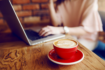Selective focus on cup of delicious coffee. In background woman using laptop.