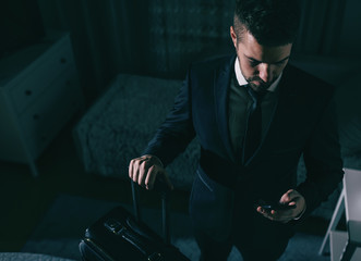Top view of businessman in suit standing in the bedroom, holding suitcase and using smart phone....
