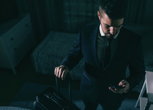 Top view of businessman in suit standing in the bedroom, holding suitcase and using smart phone. Business trip concept. - Powered by Adobe
