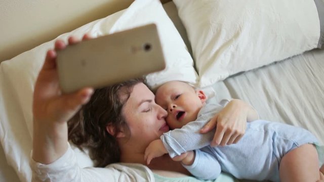 Happy mother shoots video camera phone lying in bed with her baby