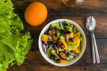 Healthy vegetarian salad with oranges, lettuce, black olives and red onion in white bowl on wooden rustic table, top view