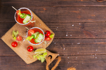 Two glasses of fresh organic tomato juice decorated with raw tomatoes, cucumber and leaves on a rustic wooden cutting board