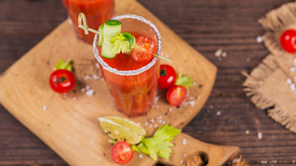 Two glasses of tomato juice decorated with fresh tomatoes, cucumber and leaves on a wooden background