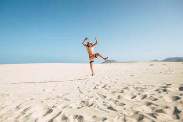 Crazy funny teenager jump with happiness on the sand of desert or beach in swimwear - young people having fun in outdoor - summer activity concept and blue sky in background