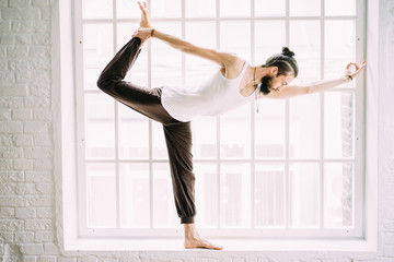 Young sporty man doing yoga exercise in white studio with wooden floor and big windows. Man and...