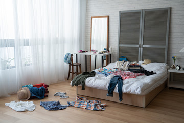 modern bright bedroom with messy clothes scatter on white bed and floor. empty room with nobody in...