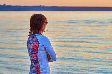 Fototapeta na wymiar A red-haired, middle-aged woman after a wake-surfing class stands on the shore of the lake and watches the sun go down.