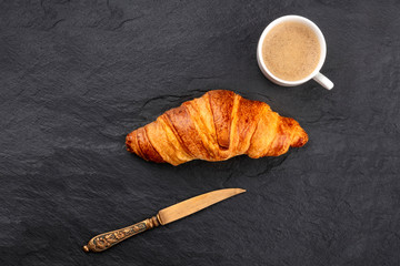A croissant with a cup of coffee and a knife, shot from the top on a black background with copy space