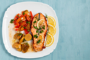 Baked salmon fillet medallion with salad of pickled vegetables and mushrooms on a white plate on a blue background, top view, flatlay, place for text, copy space