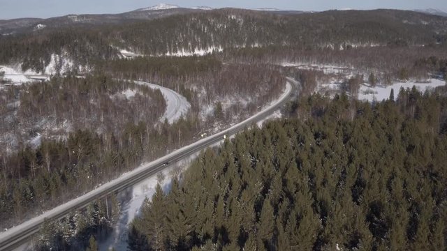 One balck wagon SUV driving along a slightly snowy asphalt freeway through a dense forest in hilly lanscapes. Winter, sunny frozen day - Aerial drone view