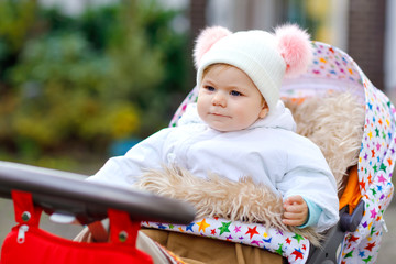 Cute little beautiful baby girl sitting in the pram or stroller on autumn day. Happy healthy child going for a walk on fresh air in warm clothes. Baby with in colorful clothes and hat with bobbles