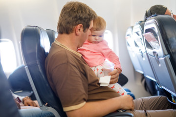 Young tired father and his crying baby daughter during flight on airplane going on vacations. Dad holding and playing with baby girl on arm. Air travel with baby, child and family concept