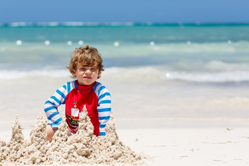 Adorable little blond kid boy having fun on tropical beach of carribean island. Excited child playing and building sand castle in sun protected swimsuit in ocean on vacations