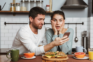 Shocked young loving couple at the kitchen have a breakfast using mobile phone.