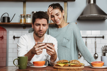 Happy young loving couple at the kitchen have a breakfast using mobile phone.