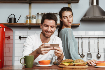 Fototapeta na wymiar Serious young woman looking at her happy man chatting by phone at the kitchen have a breakfast.