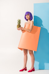 beautiful girl with purple hair covered in coral paper sheet holding pineapple while looking at camera on blue and white