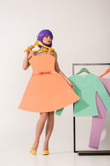 beautiful girl with purple hair holding paper dress and posing near rack with colorful clothes on white