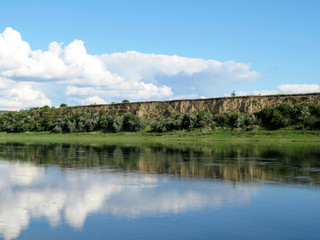 Fototapeta na wymiar Picturesque landscape with green river bank, rocky hills and blue sky. Rural scene in summer, white clouds are reflected in the water surface
