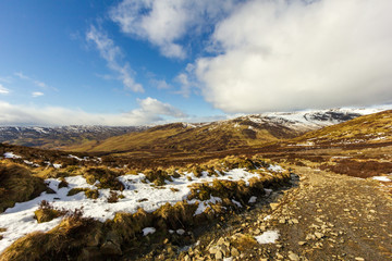 A view of a mountain path with some patchy snow under a majestic blue sky and white clouds