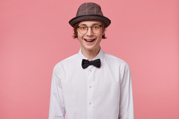 Nice young guy in white shirt, hat and black bowtie wears glasses happily widely smiling showing...