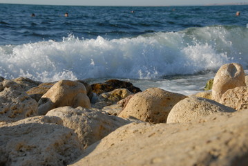 Against the backdrop of the bright blue sea, a powerful wave beating against coastal round powerful yellow boulders resembles a foaming white strip.