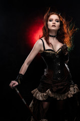 Beautiful redhead cosplayer girl wearing a Victorian-style steampunk costume with a big breast in a deep neckline  and hair blown by the wind rests on a cane. Red and black background.