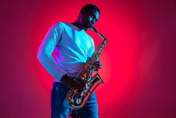 African American handsome jazz musician playing the saxophone in the studio on a neon background. Music concept. Young joyful attractive guy improvising. Close-up retro portrait. - obrazy, fototapety, plakaty