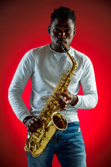 Naklejka na ściany i meble African American handsome jazz musician playing the saxophone in the studio on a neon background. Music concept. Young joyful attractive guy improvising. Close-up retro portrait.