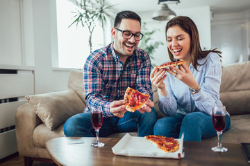 Beautiful young couple eating pizza,talking and smiling at home.