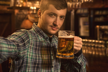 Cheerful handsome young man taking a selfie while drinking beer at the bar