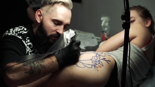 blond master corrects large peony tattoo outline with tattoo machine on young girl in gray shirt thigh lying in studio