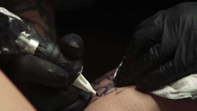 macro tattoo master in black gloves draws black outline of flower tattoo on girl thigh by professional tattoo machine