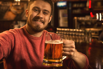 Cheerful handsome young man taking a selfie while drinking beer at the bar