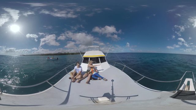 360 VR video. Parents and child sitting on the front deck of sailing yacht and enjoying water travel. They looking at ocean and swimming dolphins. Mauritius island in the distance, bright sun in the
