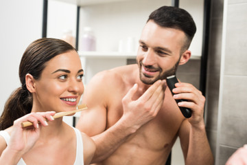 attractive woman holding toothbrush near handsome shirtless boyfriend shaving face in bathroom