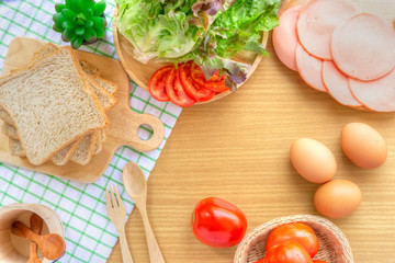 Homemade sandwich breakfast preparing. Whole wheat bread on wooden cutting board on white fabric. Slice tomatoes and lettuce on wooden plate. Fresh tomatoes, ham, eggs, spoon and fork on wooden table.