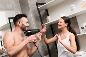 playful bearded man holding hair dryer while standing with cheerful woman in bathroom
