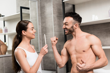 excited shirtless man gesturing while looking at cheerful girlfriend holding pregnancy test in bathroom