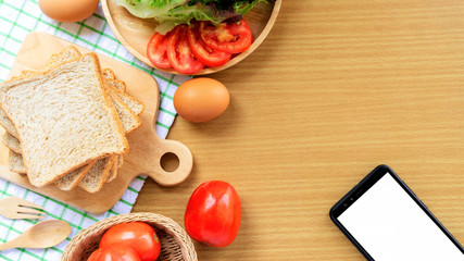 Homemade sandwich breakfast preparing. Whole wheat bread on wooden cutting board placed on a white fabric, green checkered pattern. Smart phone, slice tomatoes and lettuce placed on a wooden plate. Fr