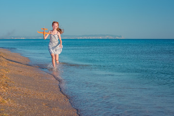 Fototapeta premium Cheerful red-haired girl playing with toy airplane and jumps over the waves of the sandy shore of the sea against the background blue summer sky. Rejoices in the rays of the sun and freedom.