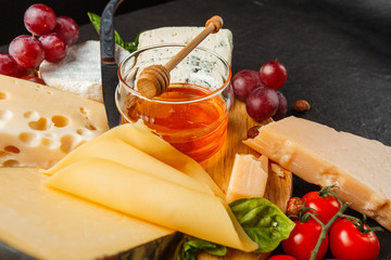 Mix cheese on dark background on wood board with grapes, honey, nuts, tomatoes and basil. Top view.