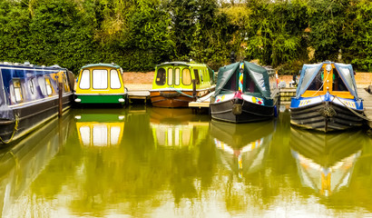 Public view of five colourful, unoccupied canal boats at their moorings in Sovereign Wharf, Banbury. Reflections of the vessels in water. Hedgerow background. Oxford canal waterway. England.