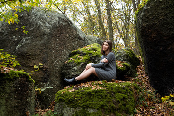 Fototapeta na wymiar A cute girl in a gray dress sits in the fall on a moss-covered stone in the forest and smiles pleasantly. Around her lie the leaves turned yellow.