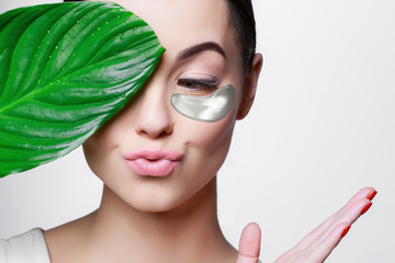 Portrait of young beautiful woman with healthy glow perfect smooth skin holds green tropical leaf. Silver cosmetics collagen hydrogel patch. Lifting anti-wrinkle mask under eyes. Skincare concept.