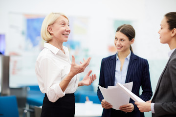 Side view portrait of smiling mature businesswoman talking to employees standing in office, copy space