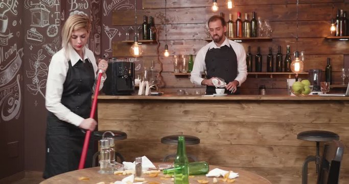 Blonde female waitress sweep the floor after a party. Cleans the table and throw the bottles away.