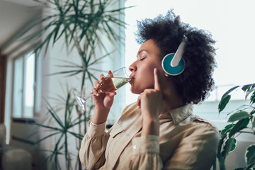 Young beautiful african american woman relaxing and listening to music using headphone, drinking wine.