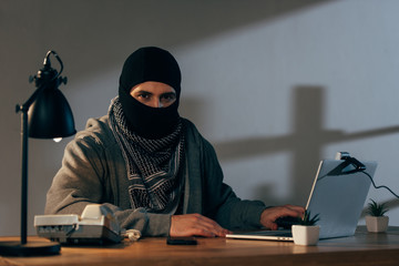 Terrorist in mask and keffiyeh scarf using laptop in room