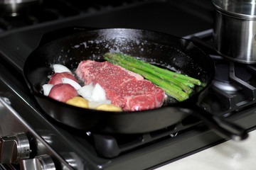 New York strip steak frying in a cast iron pan with potatoes and asparagus on a natural gas stove top.